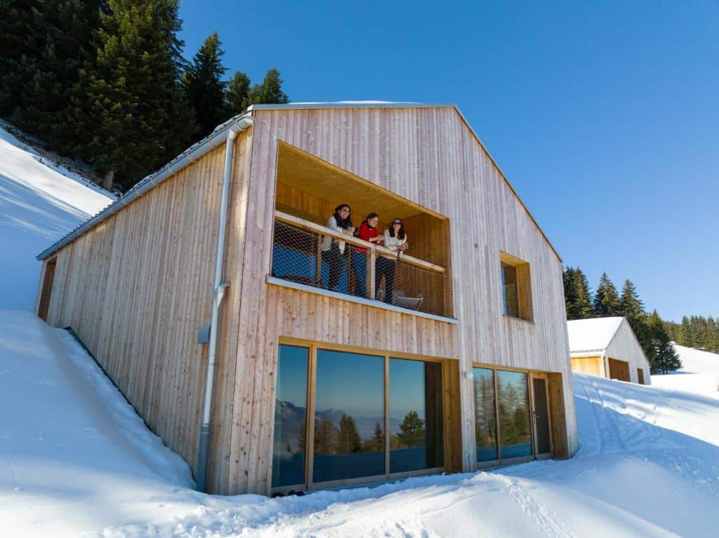 Hovedlodge Whitepod Original i snødekt landskap med besøkende på terassen og utsikt over alpene
