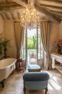 Elegant baderomssuite på Borgo Santo Pietro i Toscana med frittstående badekar, krystalllysekrone, trebjelker og utsikt mot privat terrasse og grønt landskap.