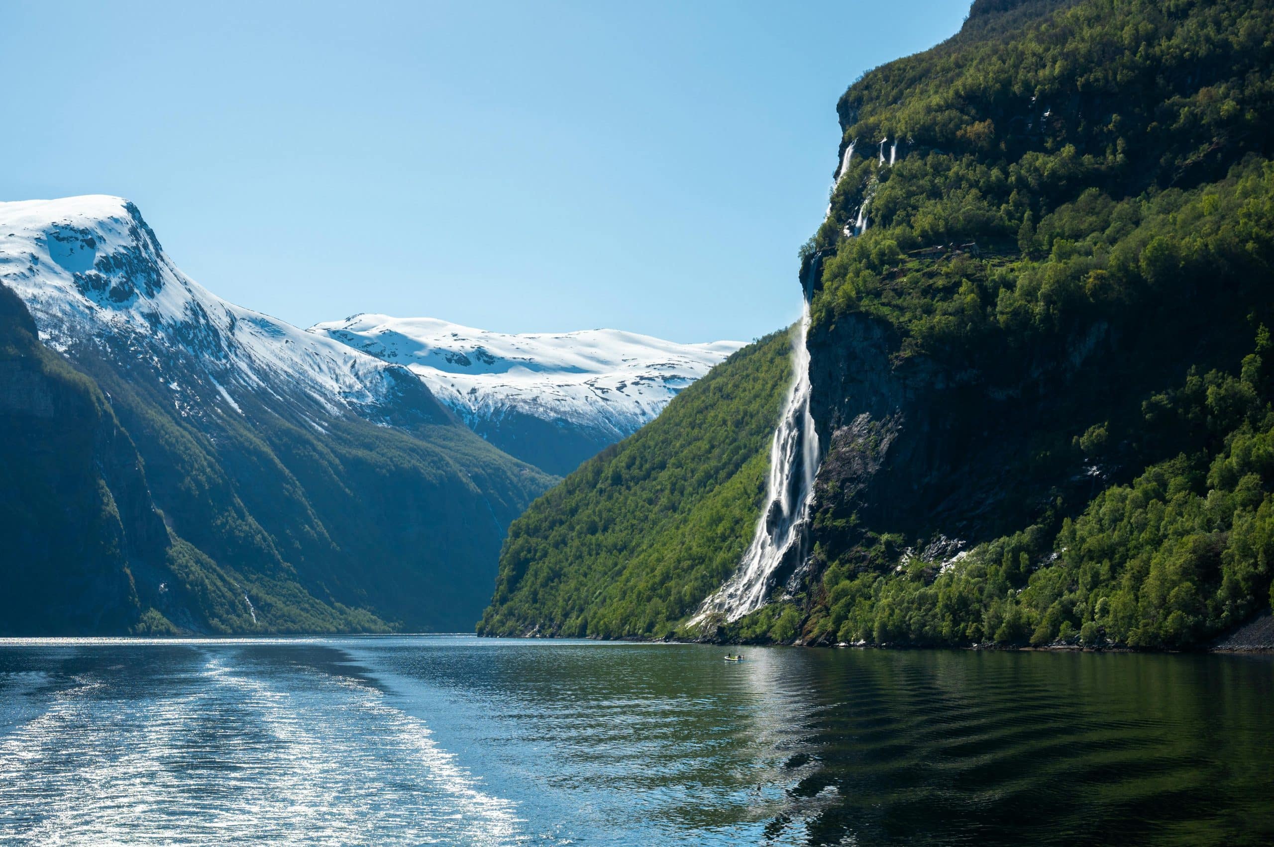 Fjellandskap i Geirangerfjorden med snødekte topper og fossefall som stuper ned i det stille, blå fjordvannet.
