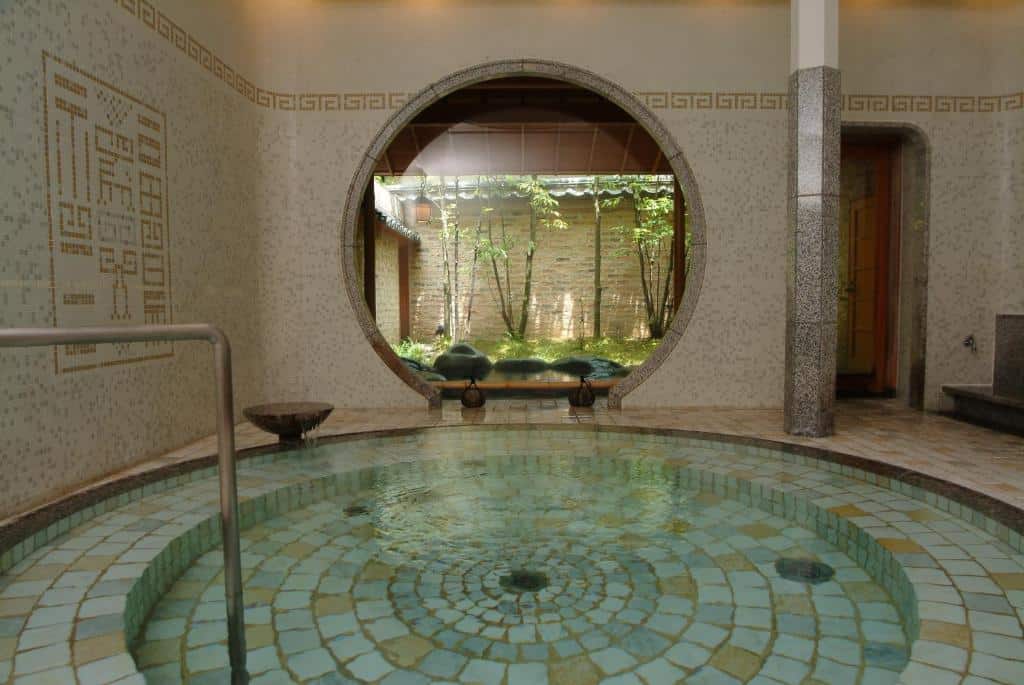 Innendørs onsen-bad med mosaikkfliser og rund åpning mot en rolig japansk hage, i et tradisjonelt ryokan-spa i Japan.