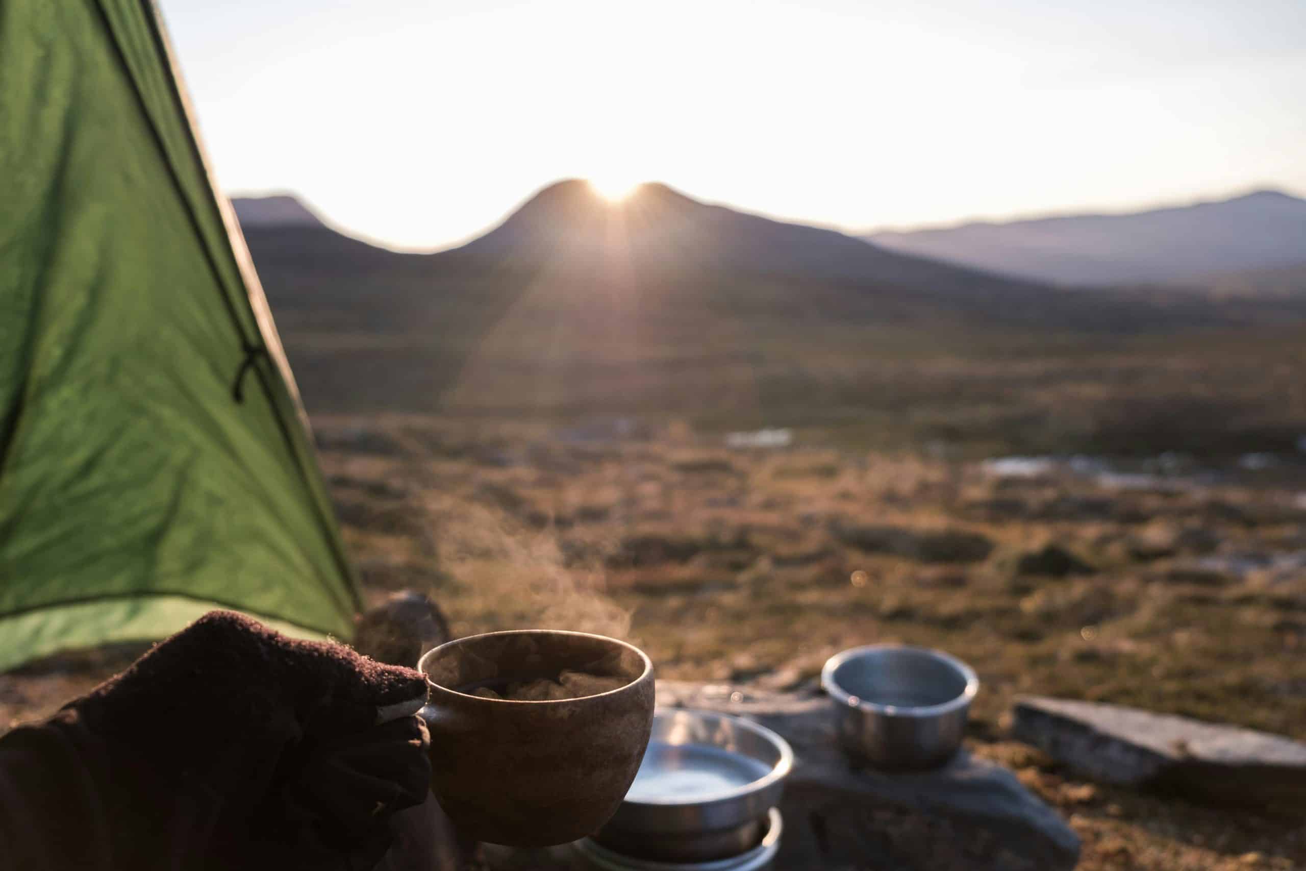 Kaffekopp ved telt i fjellet under soloppgang, med utsikt over stille landskap – rolig morgenstund på glamping- eller telttur i naturen.