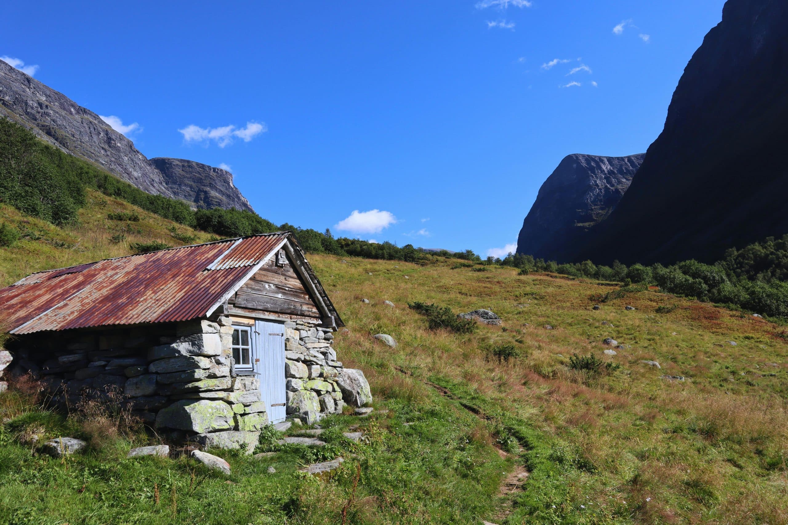 Liten steinhytte i fjellandskap nær Geirangerfjorden, omgitt av grønne enger og dramatiske fjelltopper i norsk natur.