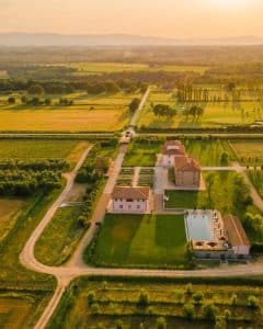Luftfoto av eksklusiv agriturismo i Toscana omgitt av vinmarker, åkre og bølgende landskap i gyllent kveldslys – landlig luksus i hjertet av regionen.