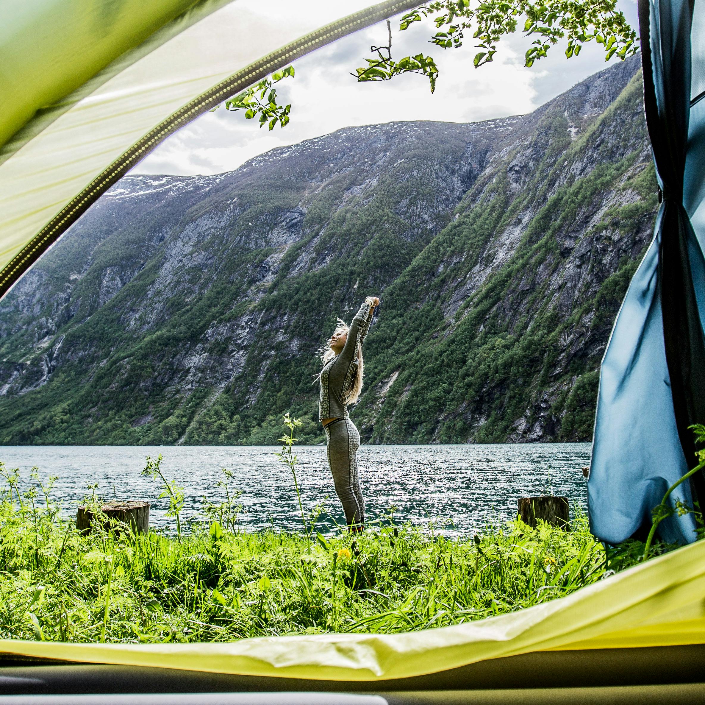 Person som strekker seg utenfor telt ved en stille fjord, omgitt av bratte fjell og grønn natur i norsk landskap.