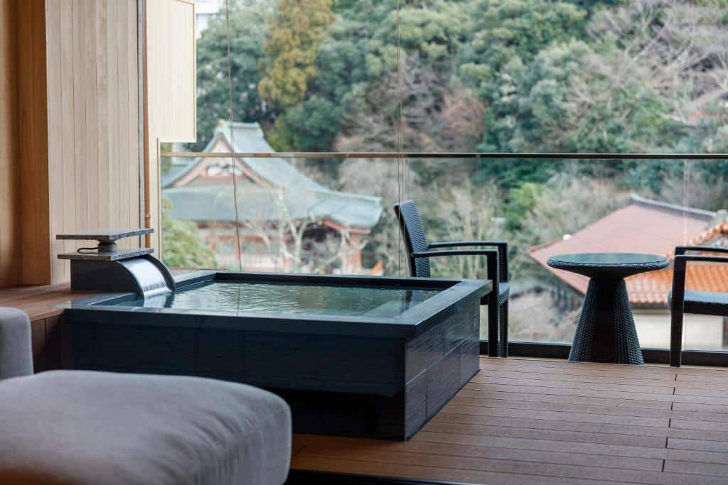 Privat onsen-badekar på balkong med utsikt mot tradisjonelt tempeltak og grønne åssider i Japan.