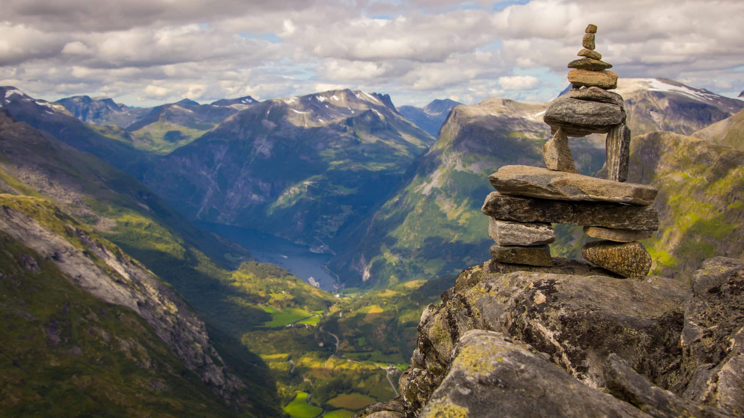 Utsikt fra fjell i Geiranger med tradisjonell steinvarde i forgrunnen og dramatisk fjordlandskap i bakgrunnen.